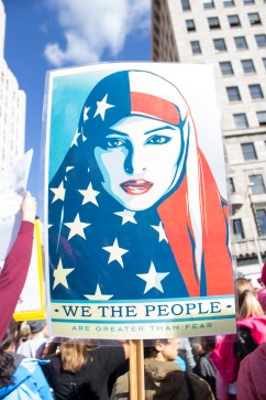 A sign held at a protest march with the drawing of a Muslim woman in an American flag hijab, a scarf the covers a woman’s hair and neck. The bottom of on the poster says We the People are Greater than Fear. This poster is a part of street artist Shepard Fairey’s We the People series designed to protest the election of Donald Trump as the American president in 2016.